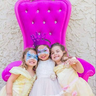 Three girls in princess dresses posing in front of pink throne chair at a children’s party for a fairytale photo setup.