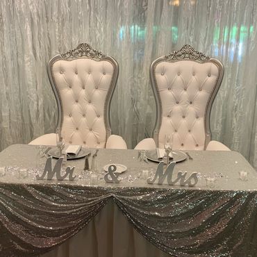 Pair of silver and white high-back thrones arranged behind glitter tablecloth for formal wedding or anniversary setup.
