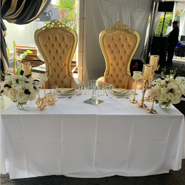 Pair of gold and champagne tufted thrones behind elegant white wedding sweetheart table with candles and roses.