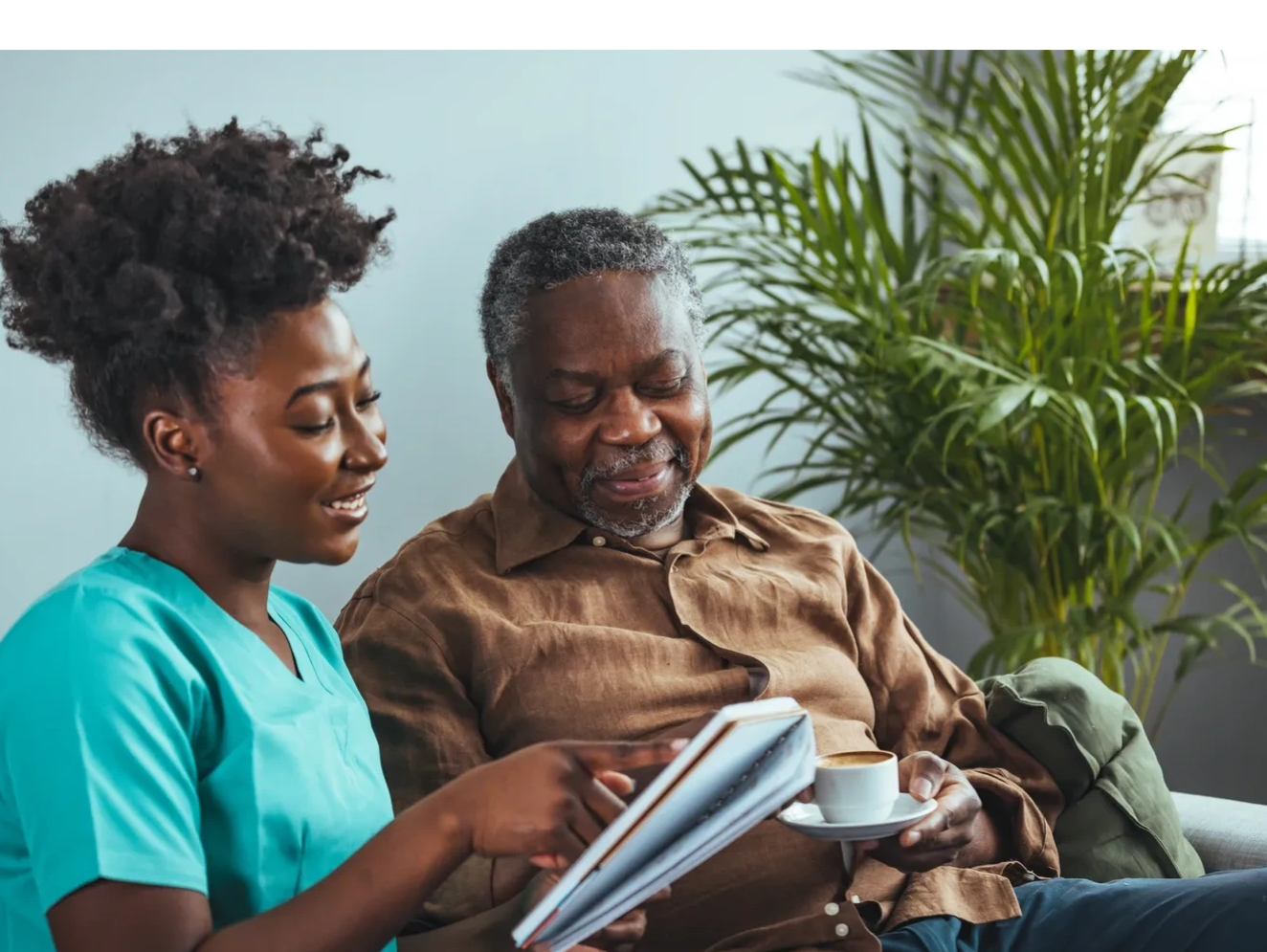 Caregiver in scrubs shows a notebook to an elderly man holding a cup of coffee.