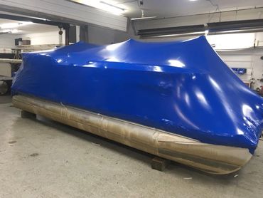 Pontoon boat covered with blue shrink wrap indoors.