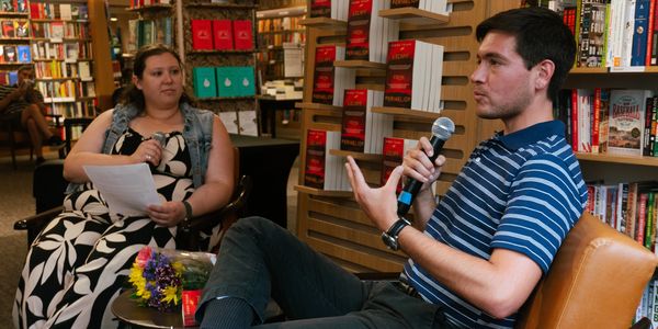 Man speaking with microphone in a bookstore interview.