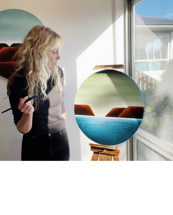 Woman painting a circular landscape on an easel near a window.