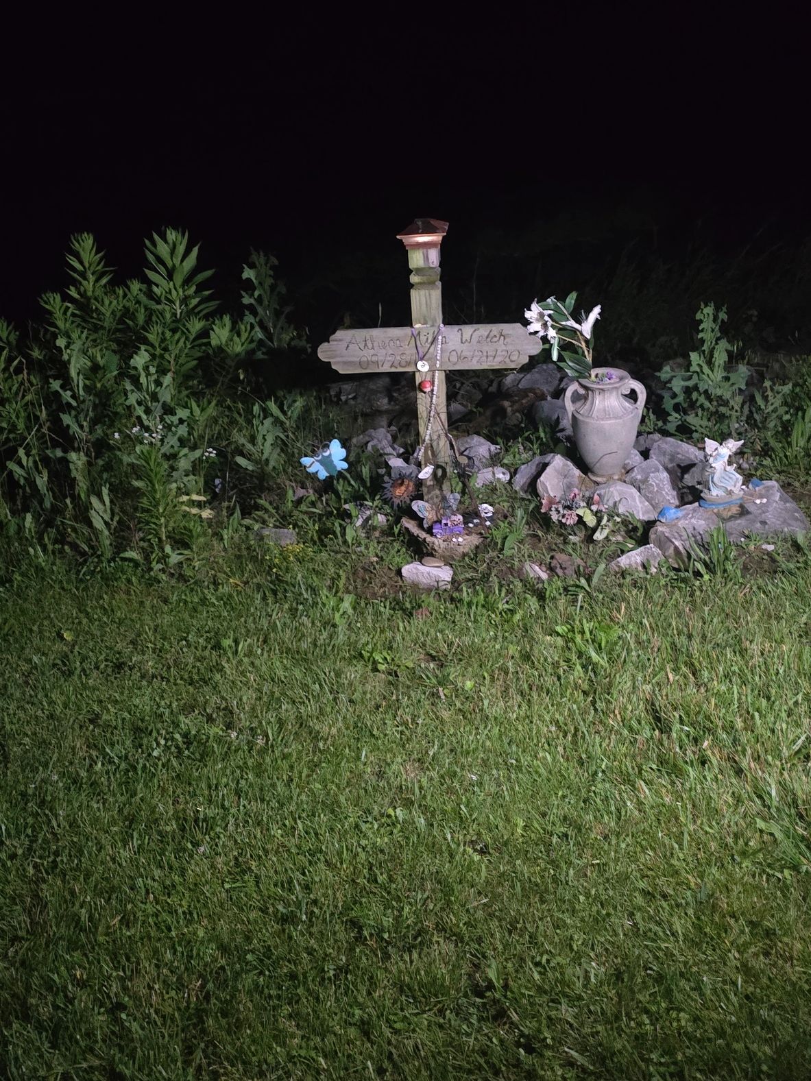 A small memorial site with a wooden cross and flowers at night.