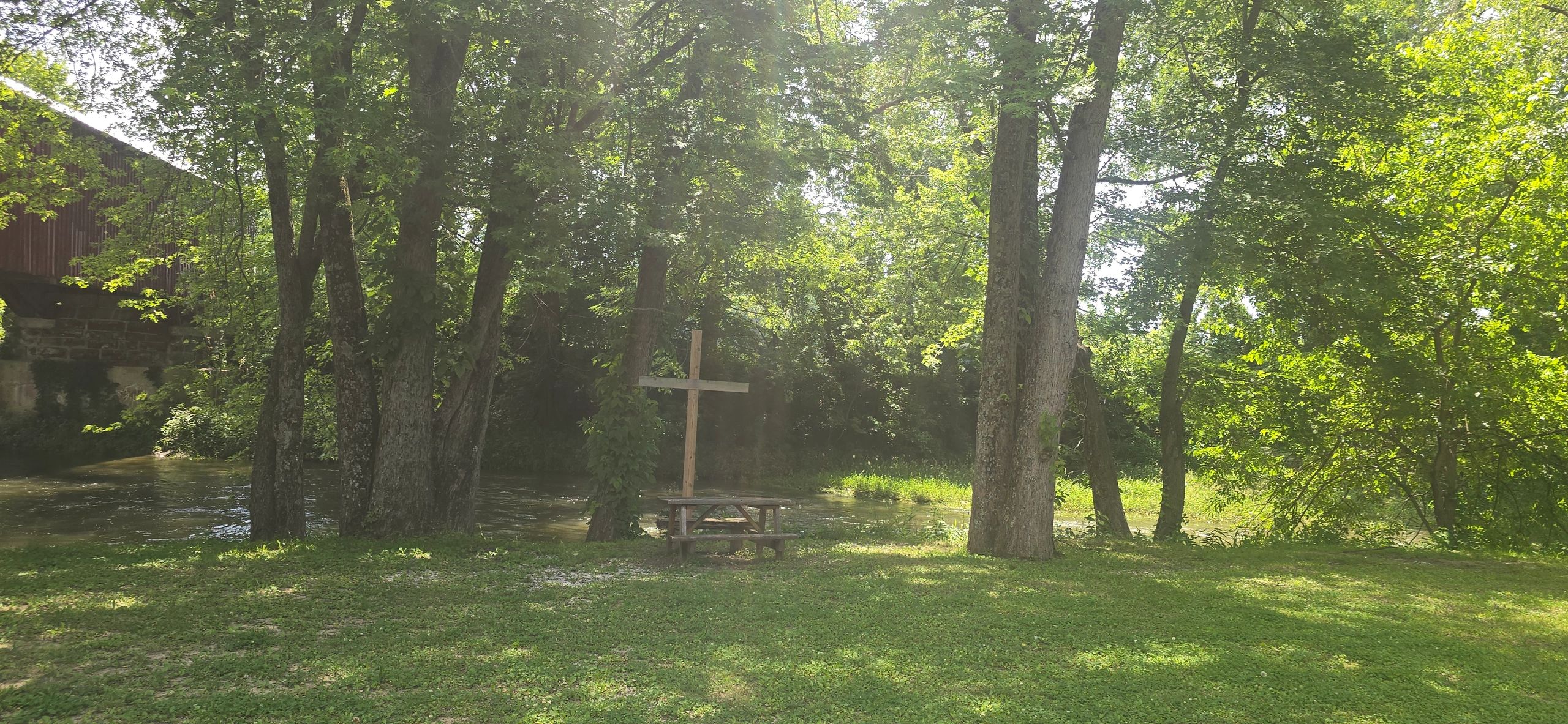 Peaceful riverside scene with a wooden cross and picnic table under tall trees.