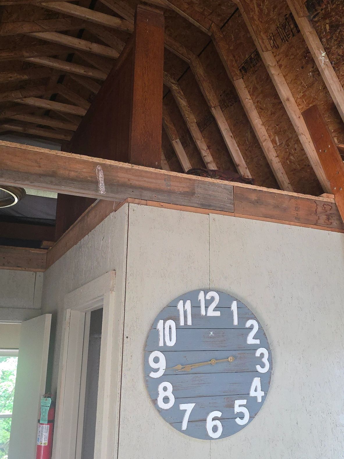 A rustic blue clock with white numbers hangs on a beige wall under a wooden loft.