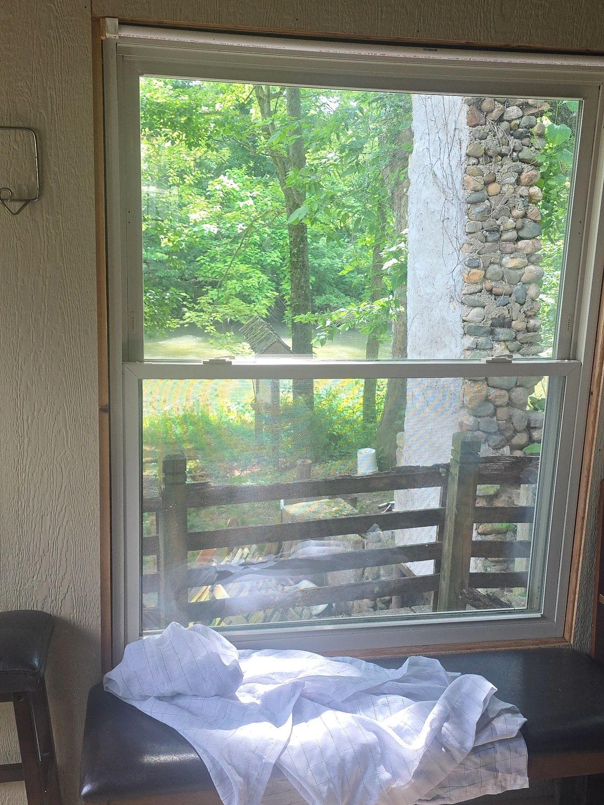 Sunlit window view of a green forest and stone chimney with a cloth on a bench.