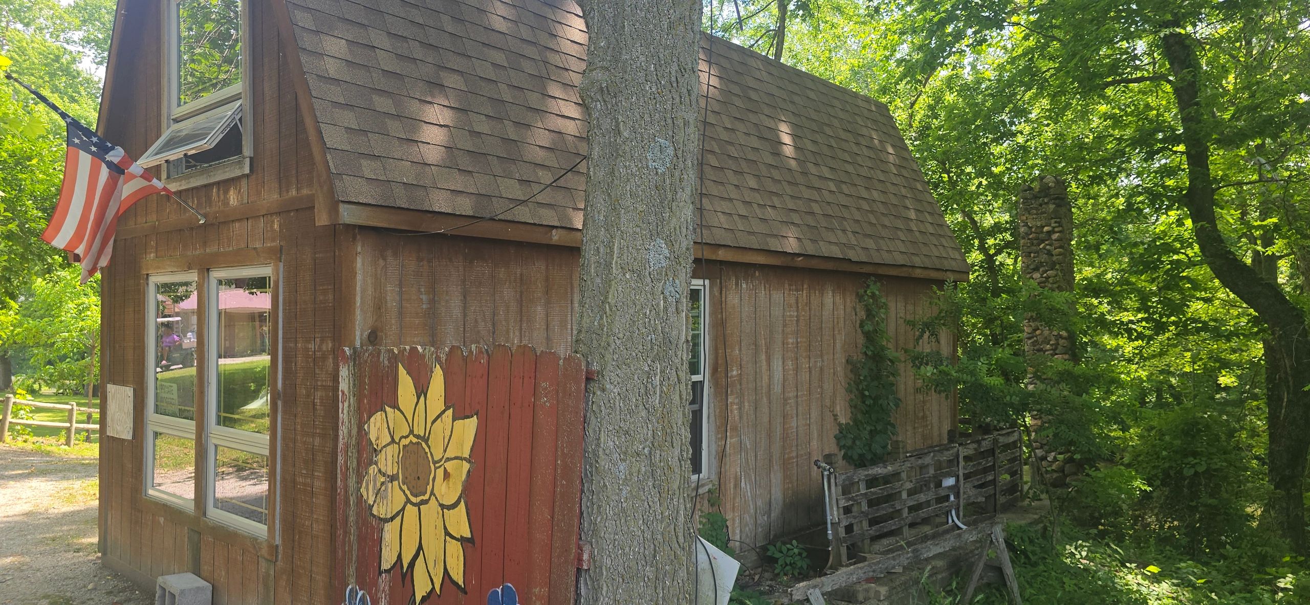 Rustic wooden cabin with an American flag and sunflower painting in a lush green forest.