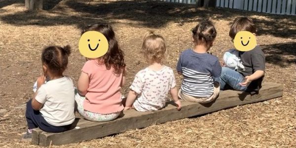 Five children sitting on a wooden beam outdoors.