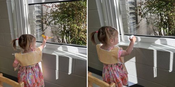 A toddler cleaning a window with different tools.
