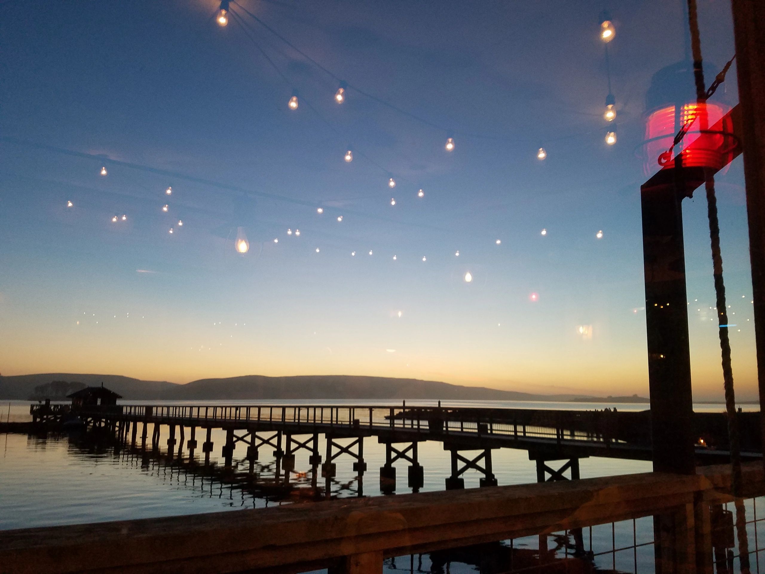 A pedestrian bridge across a calm bay at sunset in front of low lying hills, string of lights in sky