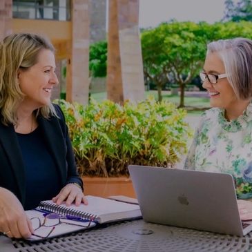 Two women smiling and chatting outdoors with a laptop and notebooks.