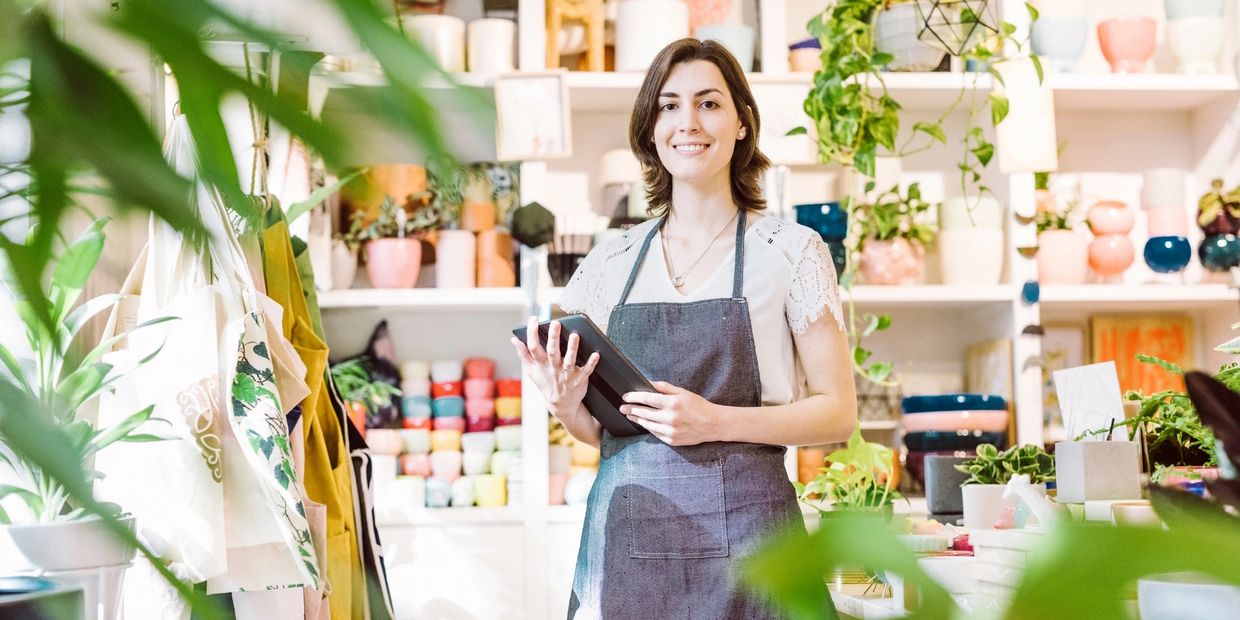 Smiling woman in apron holding tablet in a bright, plant-filled store.
