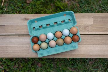 A carton of multicolored eggs on a wooden table outdoors.