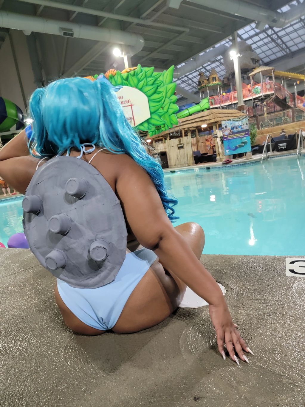 Person with blue hair and a gray turtle shell sitting by an indoor pool.
