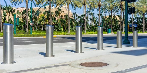 Six metal bollards on a sidewalk with palm trees in the background.