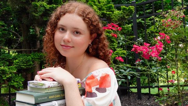 A young woman with curly red hair poses with books in a garden full of green plants and pink flowers.