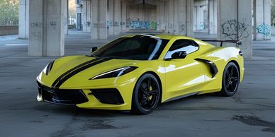 Yellow sports car with black racing stripes parked under a bridge.