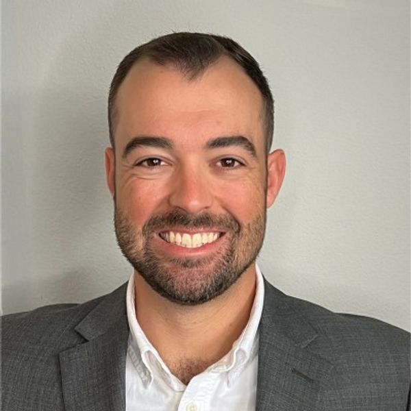 Smiling man in a gray suit jacket and white shirt against a plain wall.