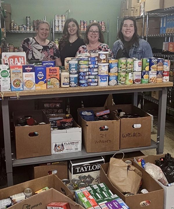 Four women standing behind a table filled with organized canned and boxed food in a food pantry.