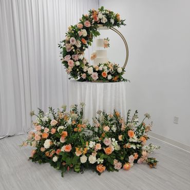 Elegant three-tier white cake surrounded by lush floral arrangements in soft peach and white tones.