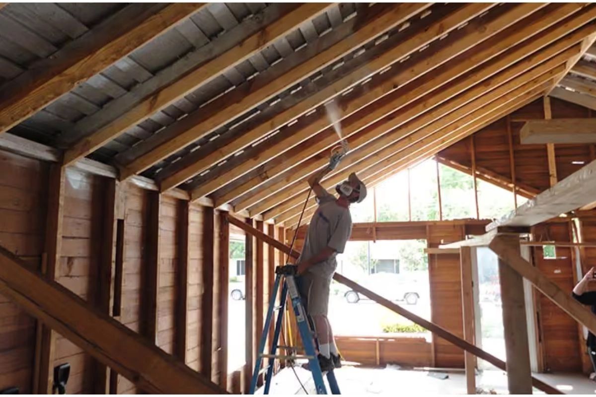 Man on ladder spraying insulation or paint on ceiling beams in wooden structure.
