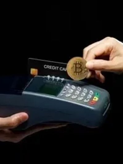 Hand holding a Bitcoin coin near a payment terminal with a credit card.