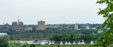 foreground: indigenous tipis at riverbank amphitheater, background: skyline brandon city
