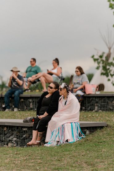 two indigenous women in regalia are focus, with people in background at outdoor amphitheater