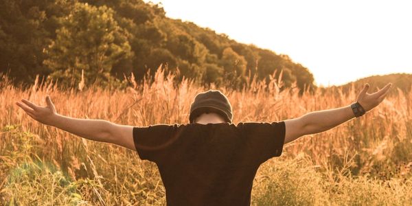 man's head bowed down, his arms outstretched, standing in sun-drenched field
