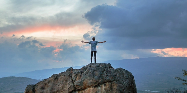 man on high cliff, standing on rock arms outstretched with clouds