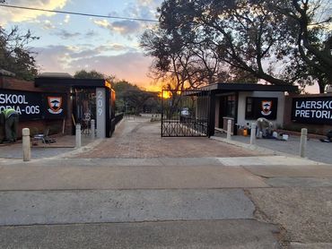 Entrance gate of Laerskool Pretoria-Oos during sunset with two workers nearby.