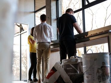 Three people discussing a project in a bright room with large windows and construction materials.