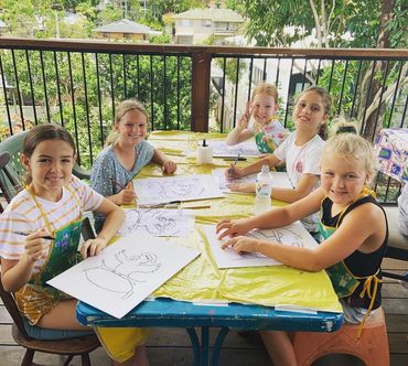 Five children happily drawing at a table outdoors with art supplies.
