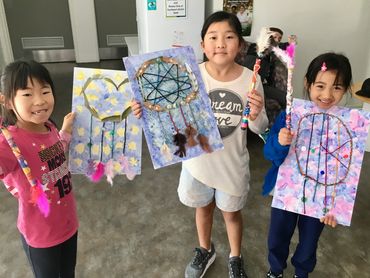 Three girls proudly show their colorful dreamcatcher crafts indoors.