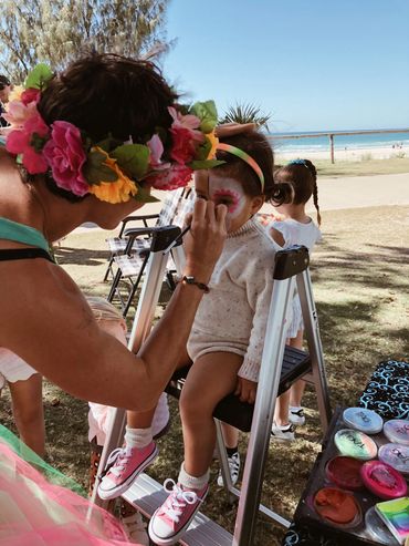 A child gets face painted outdoors near a beach, seated on a ladder.