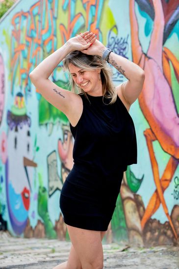 Smiling woman in black dress posing joyfully by colorful graffiti wall.