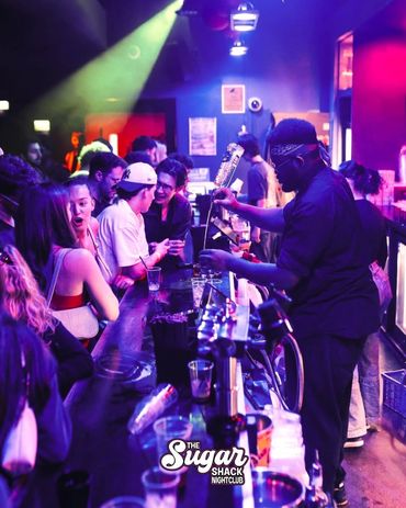 Bartender pouring drinks at a lively nightclub bar with colorful lighting.