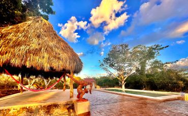Thatched roof palapa with hammock and sculpture near pool under a blue sky.