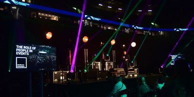 A speaker presents on stage about the role of people in events with colorful lights.