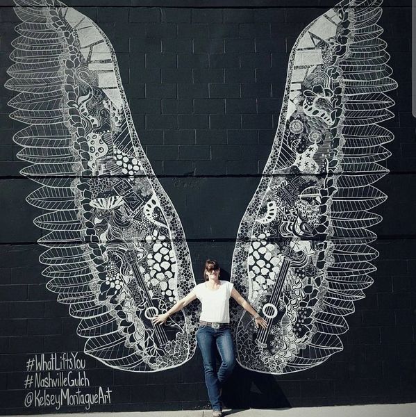 Person posing with artistic white wings mural on black wall.