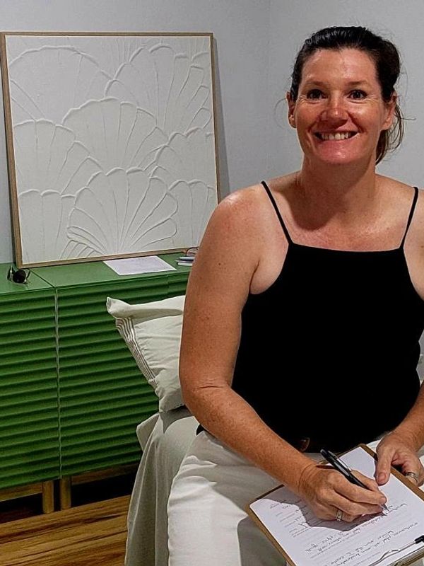 Smiling woman in a black top holding a clipboard, sitting on a bed in a modern room.