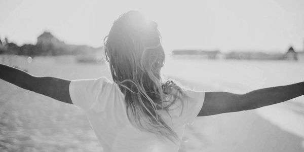 A person with outstretched arms stands on a sunlit beach, back facing the camera.