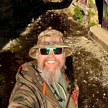 Man in camouflage gear and sunglasses smiling outdoors in sunny weather.