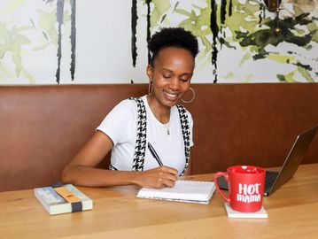 A woman smiling while writing in a notebook at a table with a laptop and a red mug.