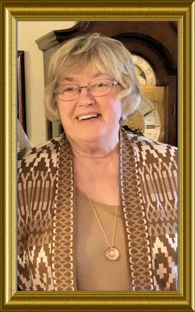 Smiling elderly woman in patterned cardigan and glasses stands in front of a grandfather clock.