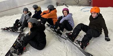 A group of six kids sitting on snow with snowboards, wearing helmets and winter clothes.