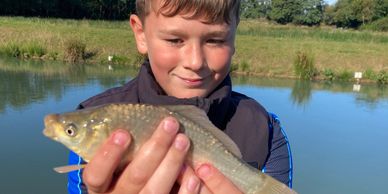 Boy proudly holding a fish by a lakeside on a sunny day.