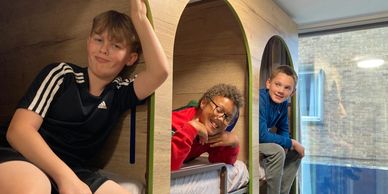 Three boys sitting inside cozy wooden pods, smiling at the camera.