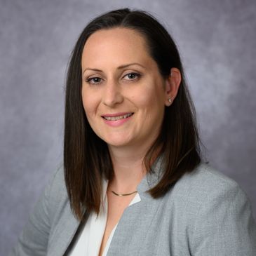 Professional woman with straight brown hair in gray blazer and white blouse smiles confidently.
